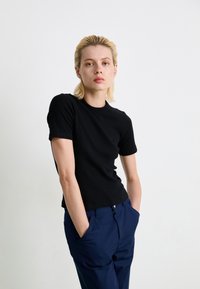 Jeune personne avec des cheveux blonds courts portant une chemise noire à manches courtes et un pantalon bleu marine, se tenant avec les mains dans les poches sur fond blanc.
