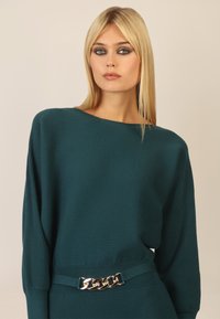 Zielona sweter o ribbowanym wzorze z szerokim dekoltem w łódkę i długimi rękawami, z ozdobnym złotym łańcuszkiem na pasku w talii.