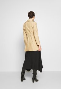 Gabardina beige con cintura ceñida, mangas largas y una apertura trasera, combinada con una falda negra fluida y botas negras hasta la rodilla.