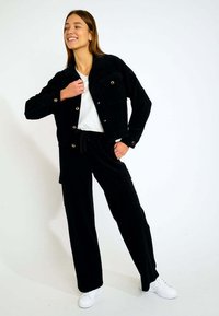 Schwarze Cordjacke mit goldenen Knöpfen und Taschen, kombiniert mit passenden schwarzen Cordhosen und weißen Sneakers. Das Outfit hat einen lässigen Schnitt.