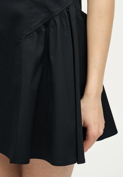 Μαύρη φούστα με πιέτες, εμφανίζεται τμήμα χεριού και μπράτσου μερικώς ορατό, σε απλό φωτεινό φόντο.