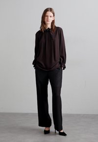 Blusa nera in satin con polsini e collo arricciati, abbinata a pantaloni neri sartoriali e décolleté neri a punta. Design semplice ed elegante.