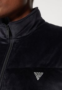 Giacca nera in velluto a zip con colletto alto e un elemento decorativo triangolare in argento sul petto, dalla texture liscia e morbida.