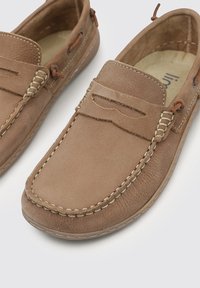 Mocasines de cuero marrón con costuras beiges, una hendidura decorativa en la parte superior y un acabado texturizado. Diseño sin cordones con perfil bajo.