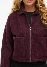 Veste courte bordeaux avec une surface texturée, un zip à l'avant et deux poches à l'avant. Dotée d'un col plat et d'une coupe ample.