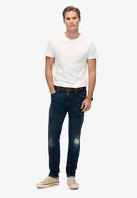 Camiseta blanca de cuello redondo, vaqueros de mezclilla azul desgastados, cinturón marrón, zapatillas en color caqui y un reloj de pulsera. Modelo de pie, con las manos en las caderas.