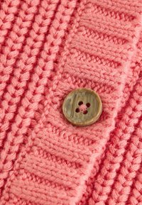 Adīta koraļļu krāsas džempera detaļa ar reljefu rakstu un lielu apaļu brūnu pogu ar četrām caurumiem.