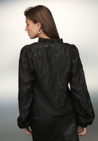 Blusa nera strutturata con maniche lunghe, scollo arricciato e motivo floreale. Indossata con una gonna in pelle, che mette in risalto dettagli intricati.