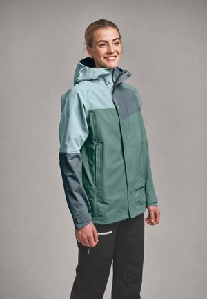 Smiling person wearing a color-blocked waterproof jacket with a hood and black outdoor pants, standing against a plain background.