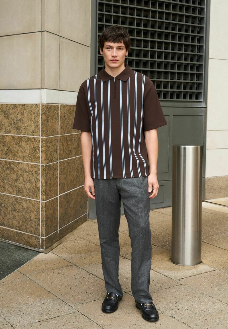 Un jeune homme se tient à l'extérieur, portant un polo marron à manches courtes avec des rayures blanches verticales, un pantalon gris et des chaussures de ville noires.