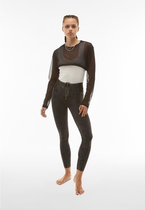 Mustaa läpinäkyvää lyhyttä toppia valkoisen tiukan tankotopin päällä, yhdistettynä korkeavyötäröisiin, tummaan farkkukankaiseen tyyliin leggings-housuihin, joissa on hienovaraisia haalistuneita kuvioita ja vetoketju.