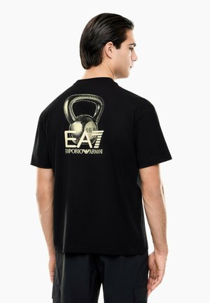 Ung mand med mørkt hår iført en sort T-shirt med en stor kettlebell-grafik og "EA7 Emporio Armani" logo på ryggen.