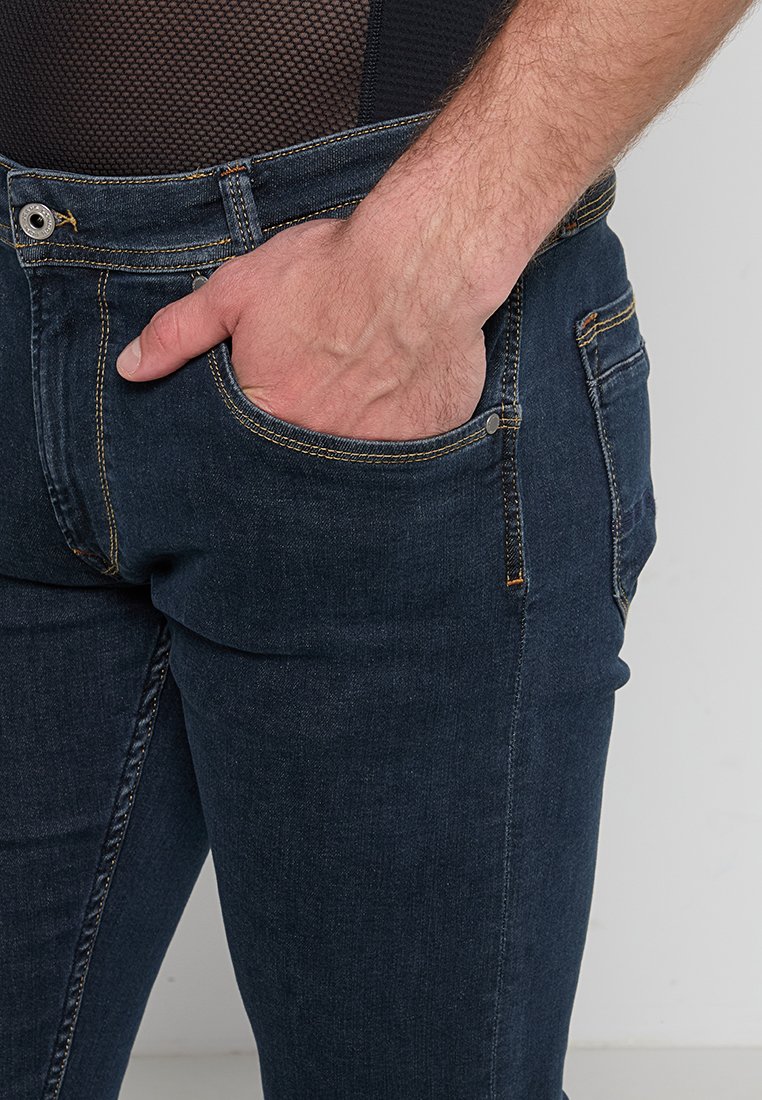Dunkelblaue Jeans werden gezeigt, mit einer Hand, die in der vorderen Tasche liegt. Der Hintergrund ist schlicht und neutral.