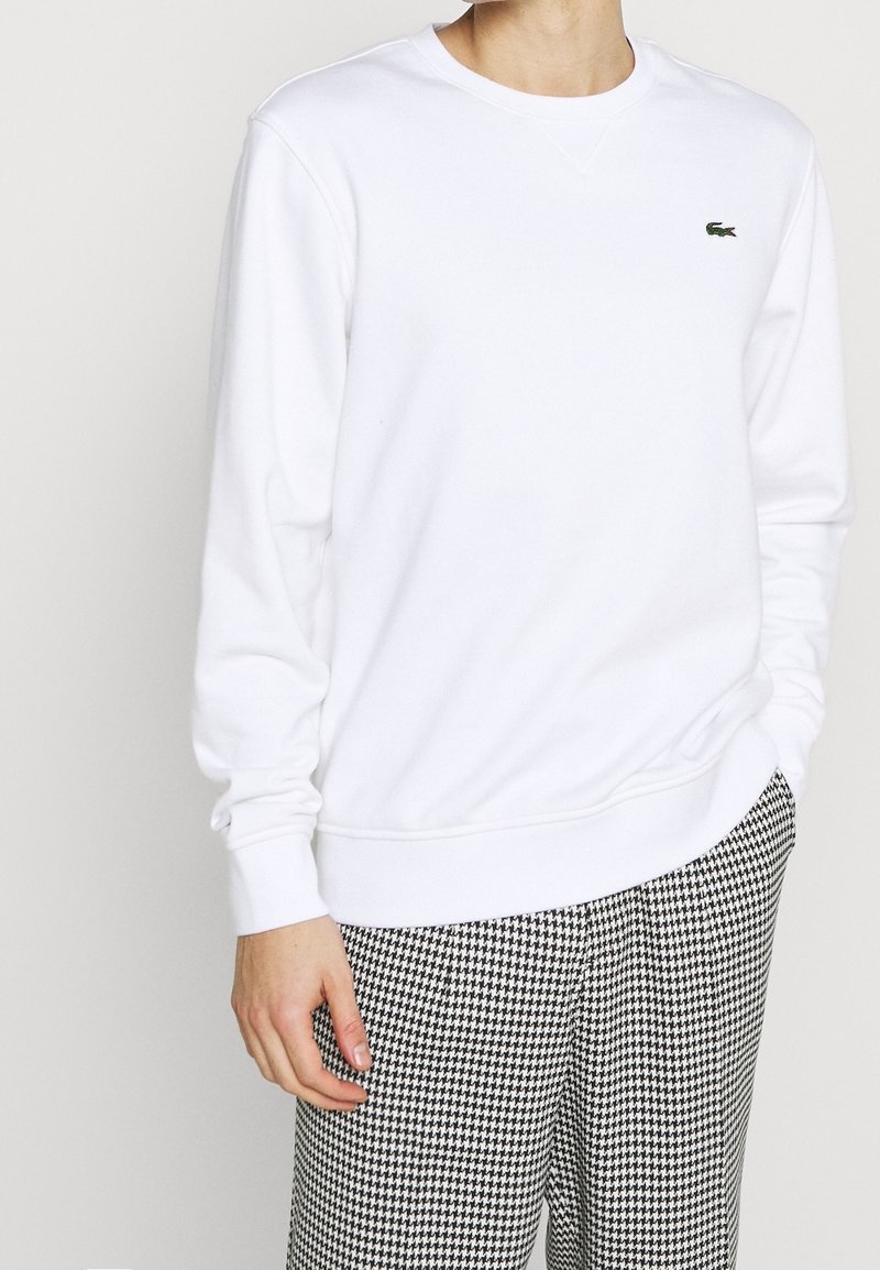 Sudadera blanca con cuello redondo y mangas largas, confeccionada en un tejido suave. Incluye un pequeño logo verde en el pecho.