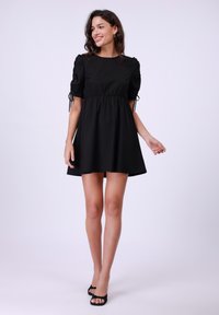 Vestido negro con mangas cortas abullonadas, cintura ajustada y lazos ajustables en los dobladillos de las mangas. Diseño de falda acampanada. Lleva sandalias negras de tacón abierto.