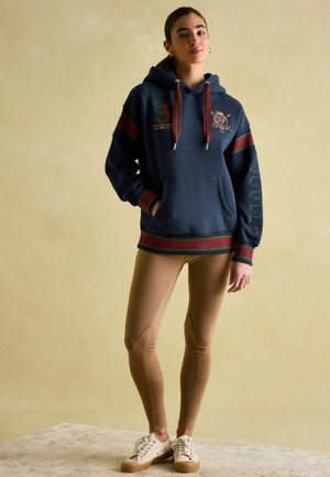 Marineblauer Hoodie mit roten und grünen Streifen, ausgestattet mit einer Kängurutasche und gestickten Details. Kombiniert mit beigen Leggings und beigen Sneakern.