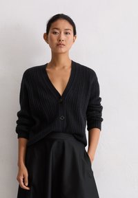 Cardigan noir en maille avec un décolleté en V profond, présentant une texture côtelée verticale, deux boutons et des manches retroussées, associé à une jupe noire.