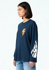 Femme aux cheveux noirs raides portant un t-shirt à manches longues bleu marine avec un éclair jaune sur la poitrine et des flammes blanches sur la manche, associé à un jean bleu.