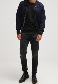 Uomo che indossa una giacca blu navy con zip sopra un maglione nero a pois, jeans scuri arrotolati e scarpe eleganti nere, in piedi contro uno sfondo semplice.