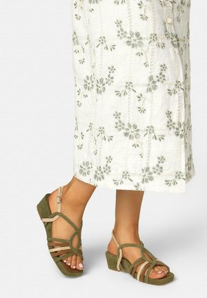 Füße in olivgrünen und beigefarbenen Sandalen mit Blockabsatz und Riemchen, stehend vor einem weißen, mit Blumen bestickten Midirock.