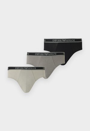 Młody mężczyzna o szczupłej, umięśnionej budowie ciała, ubrany w czarne majtki Emporio Armani, stojący boso na jednolitym jasnoszarym tle.