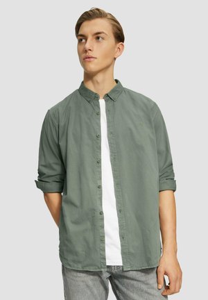 Chemise boutonnière vert clair avec col, manches retroussées, et un t-shirt blanc en dessous. Texture lisse avec un détail de logo subtil.