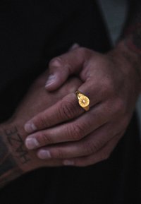 Auksinis žiedas su lapo formos dizainu, turintis centrinį brangakmenį, dėvimas ant tatuiruota ranka. Lygi tekstūra, poliruotas paviršius.