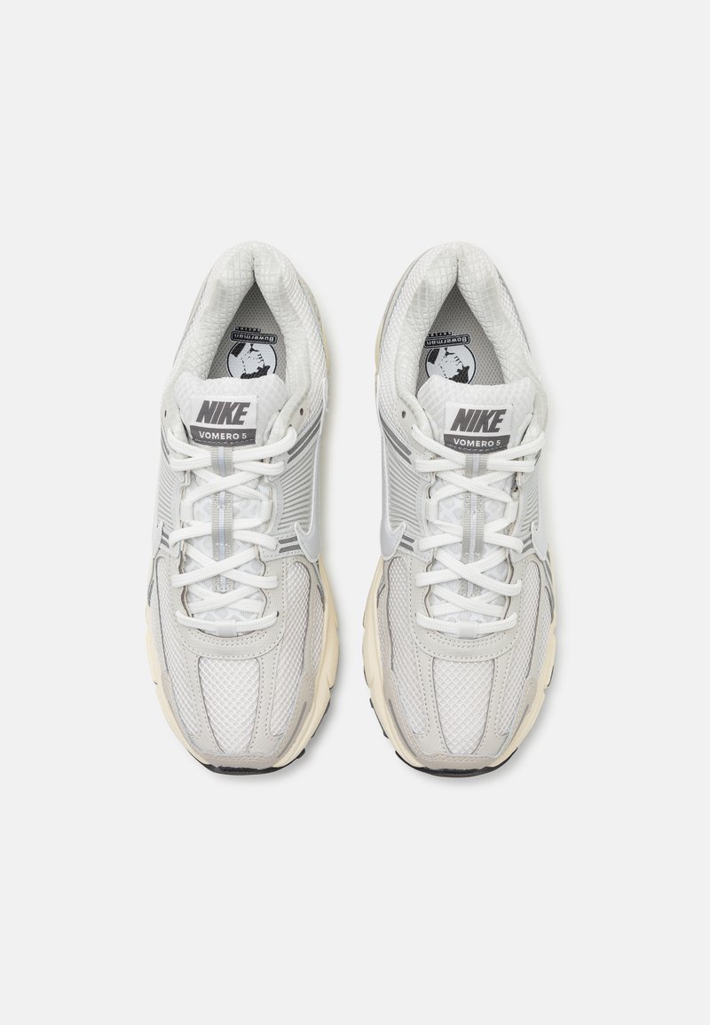 Buty Nike Vomero 5 w białej i szarej siateczce, z teksturowanymi akcentami, wyściełanym kołnierzem i białymi sznurowadłami. Gruba gumowa podeszwa z wyraźnym bieżnikiem.