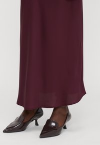 Abito bordeaux realizzato in un tessuto morbido e fluido, abbinato a tacchi a punta lucidi e scuri con un design a pattina unico.