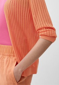 Otwarte sweter w kolorze koralowo-pomarańczowym noszony na dopasowanej różowej bluzce. Luźny krój z prążkowaną teksturą, podwiniętymi rękawami i pasującymi spodniami w kolorze brzoskwiniowym.