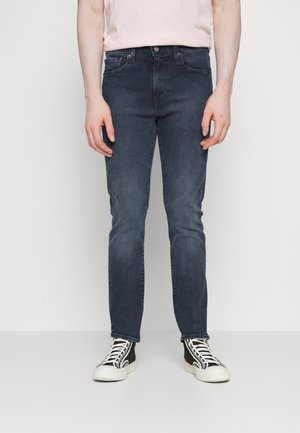 Mann trägt dunkelblaue Slim-Fit-Jeans, hellrosa T-Shirt und schwarz-weiße High-Top-Sneaker, steht vor einem weißen Hintergrund.