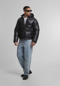 Juodas pūkinis švarkas su užtrauktuku priekyje, nuimamu gobtuvu ir elastiniais rankogaliais, derinamas su šviesiai mėlynomis džinsinėmis kelnėmis ir juodais batais.