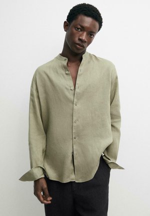 Jonge man draagt een lichtgroene, losse blouse met knopen en een opstaande kraag, staand tegen een effen witte achtergrond.