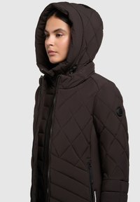 Brun quiltet jakke med høj krave og en stor hætte, som har lynlås-lukning og et logo-patch på ærmet.