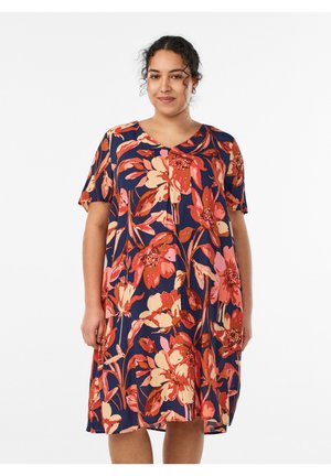 Kvinde med krøllet hår iført en knælængde marineblå kjole med store røde, orange og pink blomsterprint, stående mod en hvid baggrund.