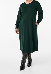 Grünes Langarmkleid mit rundem Ausschnitt und seitlichen Taschen, kombiniert mit schwarzen, genagelten Cowboystiefeln. Weicher, fließender Stoff.