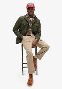 Oliwkowa kurtka z dwoma przednimi kieszeniami, koszula w kratę w odcieniach żółci i niebieskiego, beżowe spodnie i brązowe buty na sznurówki na stołku.