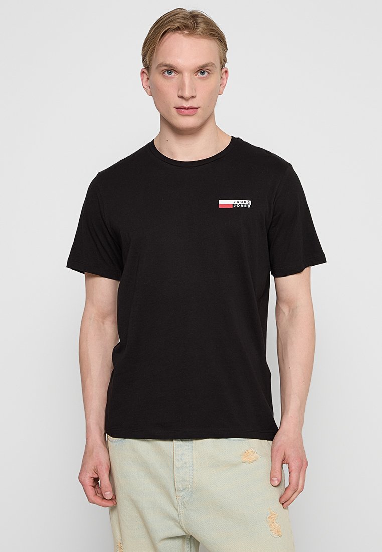 Schwarzes Baumwoll-T-Shirt mit rundem Halsausschnitt, kurzen Ärmeln und einem kleinen Grafiklogo in Rot und Weiß auf der linken Brust. Getragen mit hellen, distressierten Jeans.