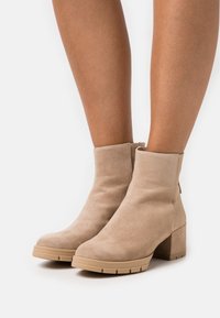 Kengät, joissa on beige mokka pinnalta, sileä rakenne, pyöreä kärki ja lohkare-korko. Takana on vetoketju ja kumipohja, jossa on urat.