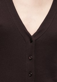 Primer plano de un cárdigan marrón oscuro con textura acanalada, tres botones negros y escote en V, mostrando la parte superior del pecho y la zona del cuello.