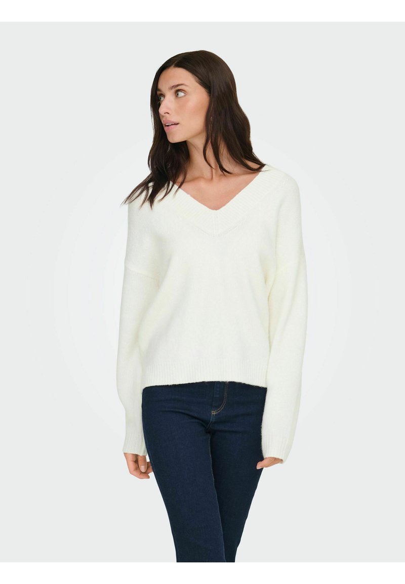 Pull blanc en maille avec col en V, coupe décontractée, poignets et ourlet côtelés, associé à un jean bleu foncé. Texture lisse, design minimaliste.