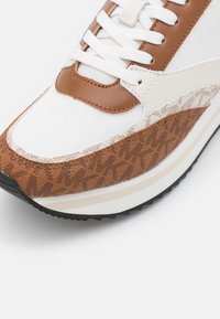 Valkoinen lenkkari, jossa ruskeat nahkaiset yksityiskohdat, teksturoitu ruskea kuvioitu paneeli ja kontrastinen beige välipohja. Nauhat klassisessa tyylissä.