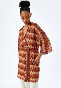 Lång kimono-liknande rock i röd, orange och guld zigzag-mönster. Lätt vikt, V-hals, vida ärmar och ett bälte att knyta i midjan.
