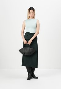 Černá kabelka s křivkovou siluetou, kterou nosí modelka v světlém zeleném topu a tmavě zelené sukni, doplněná dlouhými černými botami.