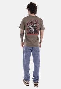 Uomo con capelli ricci, tatuaggi, indossa una maglietta marrone con grafica di un cane e la scritta "Propaganda", jeans azzurri e sneakers marroni Nike, e si trova di spalle.