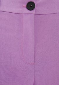 Levandulové kalhoty s hladkou texturou, které mají černý knoflík a růžové prošívání na švu postranní kapsy.