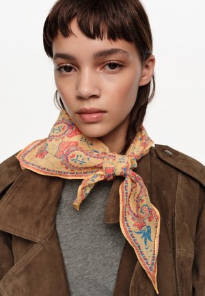 Joven con cabello corto castaño, que lleva una chaqueta de ante marrón y una bufanda amarilla con estampado atada alrededor del cuello.