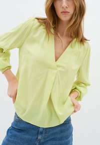 Blusa verde chiaro con uno scollo a V profondo, maniche morbide, polsini elastici e vestibilità comoda. Realizzata in un tessuto morbido e liscio.