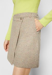 Jupe en tweed beige avec des éclats multicolores. Présente une poche latérale, un bouton près de la taille et une forme légèrement évasée.
