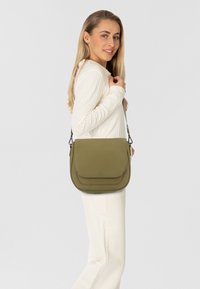 Donna con lunghi capelli biondi, vestita con un outfit bianco, mentre porta una borsa a tracolla di colore verde oliva con patta sulla spalla, sorridendo leggermente.